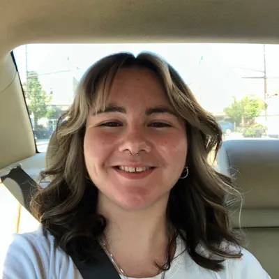A young woman with shoulder-length wavy brown hair and bangs, featuring a double nose piercing and a silver hoop earring, smiles broadly with a toothy grin while sitting in the passenger seat of a car, wearing a white shirt and a seatbelt.