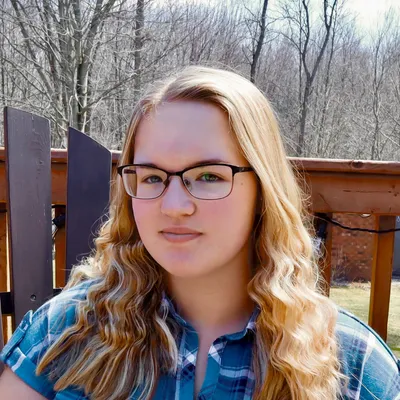 A young woman with long, wavy blonde hair and rectangular glasses, wearing a blue and white plaid shirt, smiles softly while sitting outdoors on a wooden deck with trees in the background.