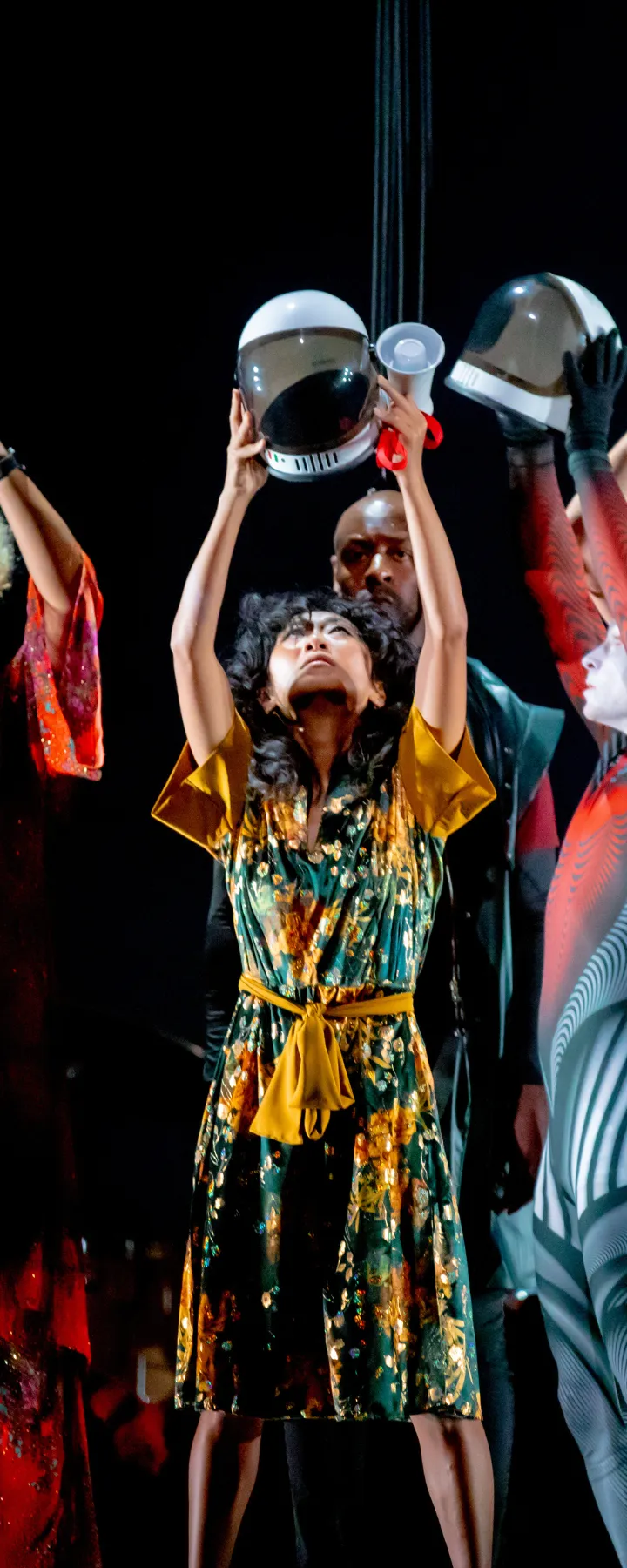 Five performers in colorful costumes hold bowls above their heads on stage, creating a dynamic and vibrant scene.