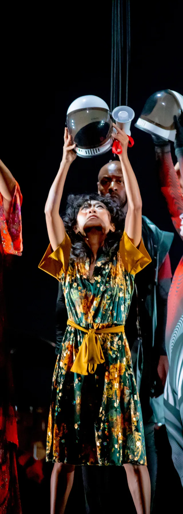 Five performers in colorful costumes hold bowls above their heads on stage, creating a dynamic and vibrant scene.