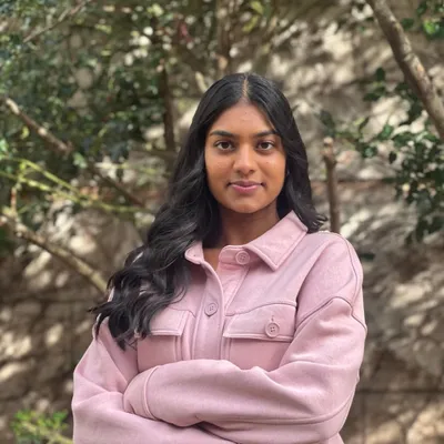 A young woman with long, wavy dark hair parted in the middle gives a subtle, confident smile with her arms crossed, wearing a light pink button-up jacket outdoors in front of blurred green foliage and a stone wall.