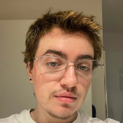A young man with brown hair, blue eyes, wire-rimmed glasses, a small chin goatee, and a black stud earring, looking directly into a mirror with a neutral expression.