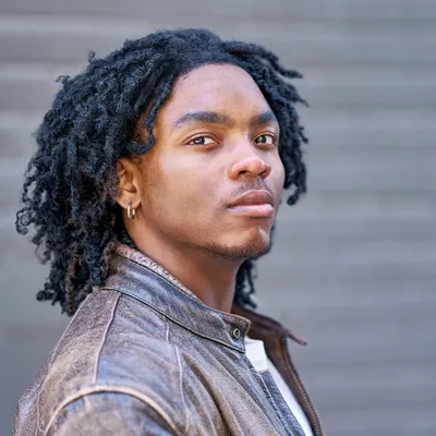 A portrait of a young man with dark twisted locs and a small goatee, wearing a silver hoop earring and a textured brown leather jacket over a white shirt, looking calmly off-camera against a blurred grey wooden fence.