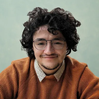 A young man with voluminous, curly dark hair, thin wire-rimmed glasses, and a goatee gives a gentle, closed-mouth smile while wearing a textured burnt-orange sweater layered over a white collared shirt with a small polka-dot pattern, set against a plain, muted sage-green background.