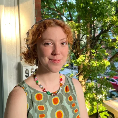 A young woman with curly reddish hair held back by a dark headband, wearing a colorful beaded necklace and a sleeveless sage-green top with a bold orange and yellow circular pattern, smiles gently while standing outdoors next to a building with the number "95" visible, under the shade of a leafy tree.
