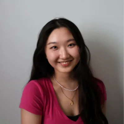 A smiling young girl with long dark hair wearing a pink top and layered necklaces, posing against a plain white background.