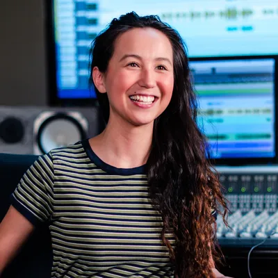 A woman with long, textured dark hair and a radiant, open-mouthed smile, wearing a horizontally striped navy and green t-shirt, sits happily in a recording studio surrounded by computer monitors and an audio mixing console.