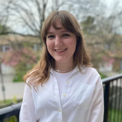 A young woman with light brown hair featuring bangs smiles warmly, showing her teeth, while wearing a white button-down shirt over a white top and a silver chain necklace, standing outdoors on a balcony with blurred early spring trees and a black railing in the background.