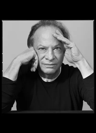 A black-and-white portrait of an older man with a prominent forehead and wispy hair, looking directly at the camera while resting his face against his hand and touching his forehead with the fingers of his other hand.