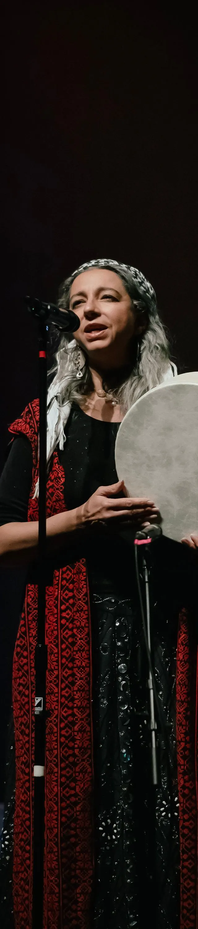 A woman with long gray hair, wearing a black outfit adorned with red patterned trim, stands on a dark stage singing into a microphone while holding a large, circular frame drum.