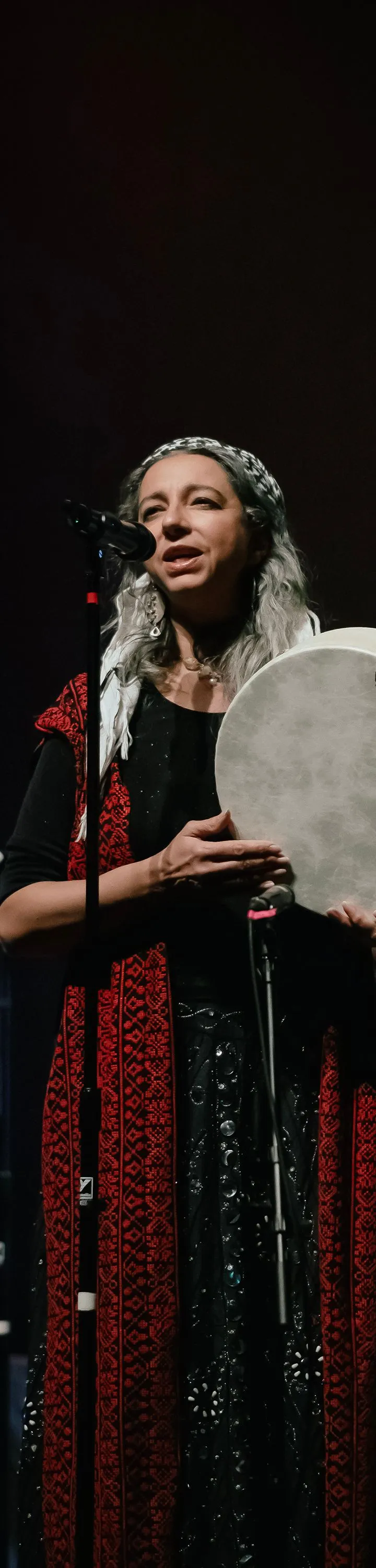 A woman with long gray hair, wearing a black outfit adorned with red patterned trim, stands on a dark stage singing into a microphone while holding a large, circular frame drum.