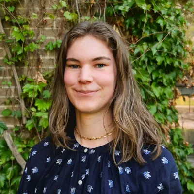 A young person with shoulder-length light brown hair and blue-green eyes gives a soft, closed-mouth smile while wearing a gold link chain necklace and a dark blue blouse with a light blue floral print, standing outdoors in front of a brick wall covered in green ivy.