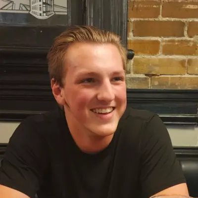 A young man with light brown hair styled back and a broad, joyful smile, wearing a plain black t-shirt, is pictured indoors looking slightly off-camera in front of a rustic yellow brick wall and dark window frame.