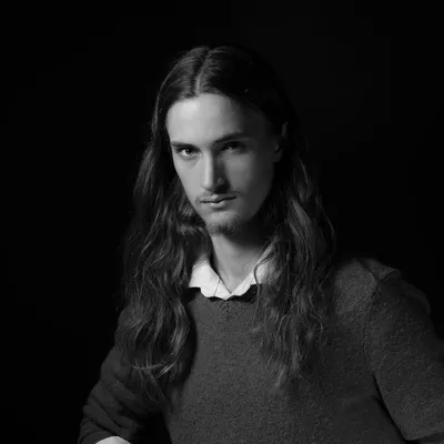 A dramatic black-and-white portrait of a long-haired young man with a light goatee, looking directly at the camera with a serious expression while wearing a dark knit sweater over a white collared shirt, set against a completely black background.