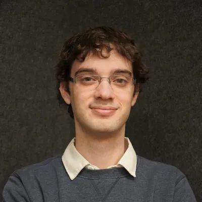 A young man with glasses and short, curly brown hair smiles at the camera. He is wearing a gray sweater over a white shirt, and standing against a neutral dark gray background.
