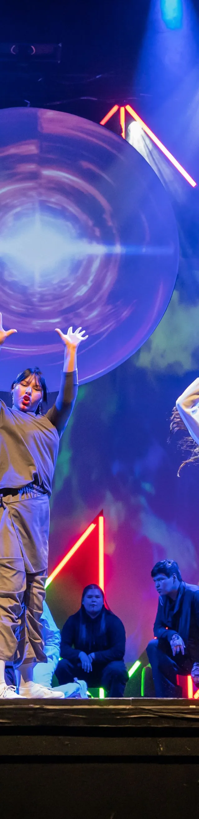 A large group of dancers, primarily in grey, black, and white outfits, perform a synchronized routine on a brightly lit stage, with several front-row members raising their arms and hands while a large circular projection and colorful neon lights illuminate the backdrop.