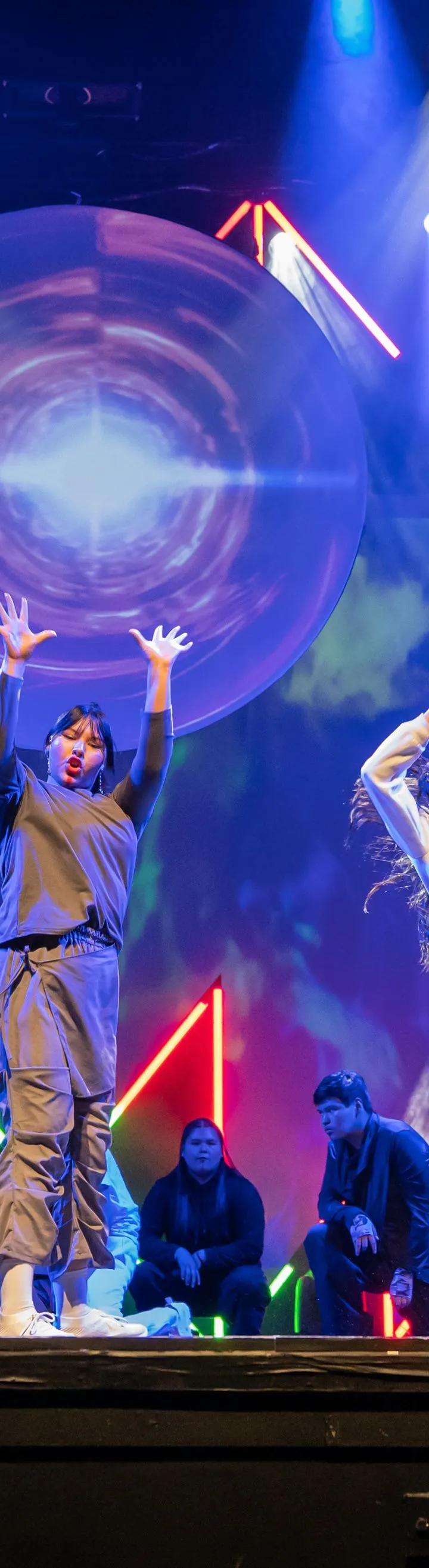 A large group of dancers, primarily in grey, black, and white outfits, perform a synchronized routine on a brightly lit stage, with several front-row members raising their arms and hands while a large circular projection and colorful neon lights illuminate the backdrop.