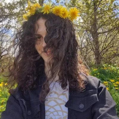 A young person with long, voluminous dark curly hair, partially obscuring one eye, wears a crown of bright yellow dandelions while smiling gently outdoors in a grassy, sunlit field.
