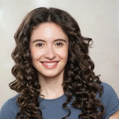 A woman with dark, voluminous spiral curls and a bright, wide smile showing her teeth, wearing a textured blue crewneck top, poses directly facing the camera against a plain, neutral background.