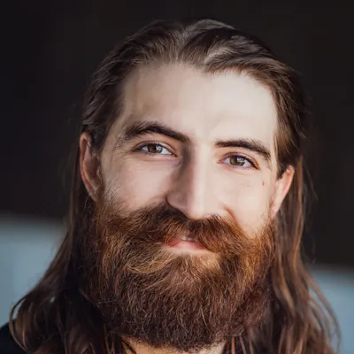 A close-up portrait of a smiling man with long dark hair and a thick, reddish-brown beard, set against a dark, softly lit background.