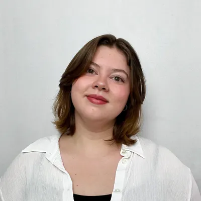 A young woman with a warm smile, brown hair parted in the middle, and red lipstick, wearing a white collared shirt over a black top against a plain light background.