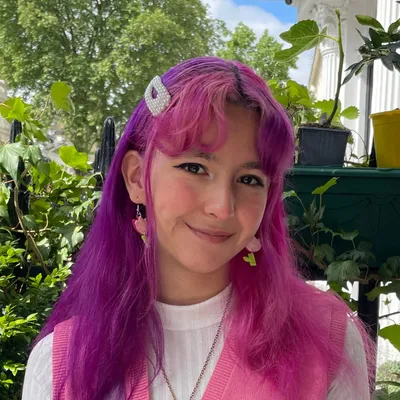 A young woman with vibrant two-toned purple and magenta hair, styled with a pearl-encrusted hair clip and face-framing bangs, gives a gentle, closed-mouth smile while standing outdoors among lush green leaves, wearing a white shirt layered under a pink vest.
