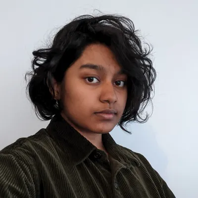 A young person with dark, thick, wavy hair and a neutral, calm expression poses against a plain white wall wearing an olive-green corduroy button-up shirt.