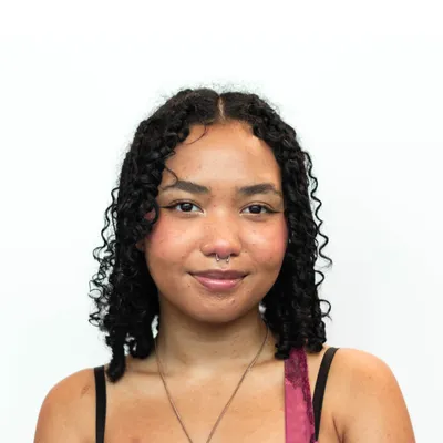A young woman with shoulder-length, dark curly hair and a gentle smile, wearing a silver septum ring, black winged eyeliner, and a thin silver chain necklace, poses against a plain white background with a black strap and a glimpse of a magenta lace strap visible on her shoulders.