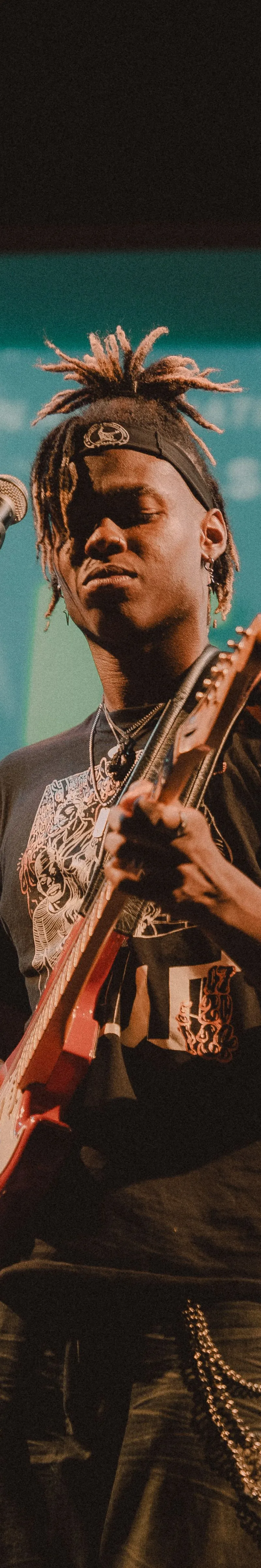A musician with dreadlocks tied up in a headband stands on stage, playing a red electric guitar while singing into a microphone, set against a teal backdrop with large, partially visible white text.