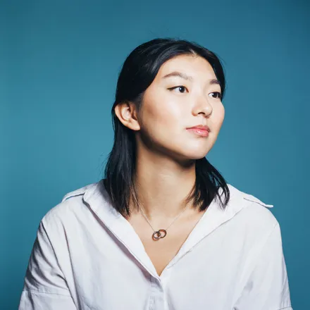 A woman with black shoulder-length hair, wearing a white shirt and a simple necklace. She looks away from the camera, to the right, set against a blue background.