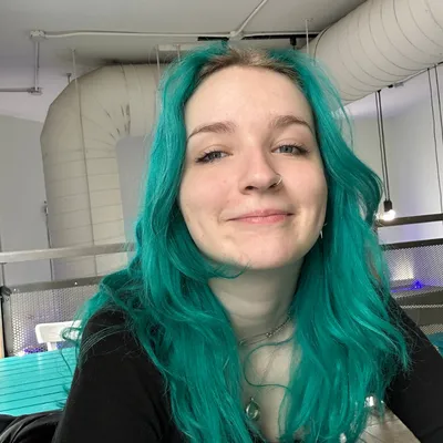 Young person with bright teal hair and a nose ring smiles warmly in an industrial setting with exposed pipes and lights.