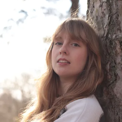 A young woman with light brown hair leans against a tree outdoors, softly lit by sunlight with a blurred natural background.