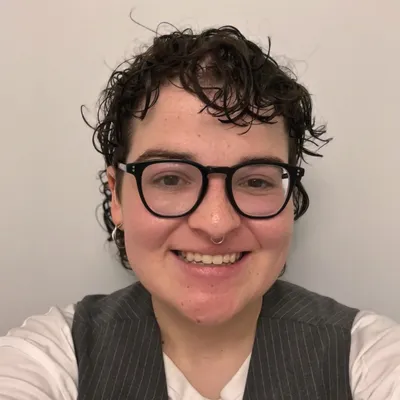 A smiling person with curly hair and glasses, wearing a gray vest over a white shirt. Neutral background. Expresses a friendly, casual vibe.