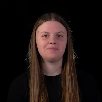A young person with long, straight light-brown hair and a neutral, slightly pursed-lip expression, wearing a simple black top, is captured head-on against a pitch-black background.