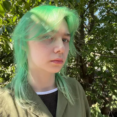 A young person with vibrant, shoulder-length mint-green hair and a neutral expression, wearing an olive-green jacket over a black mesh and white top, is captured outdoors in dappled sunlight surrounded by lush green foliage.