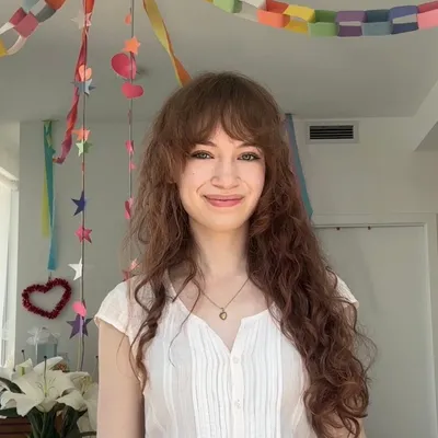 A smiling young woman with long, wavy reddish-brown hair and bangs, illuminated by warm, bright backlight outdoors.