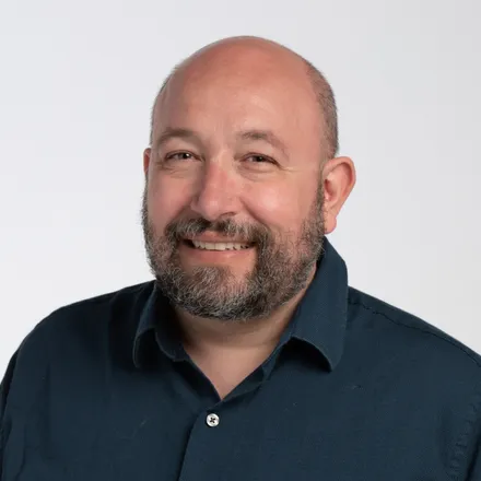 A bald man with a beard smiles warmly in a portrait against a plain white background, wearing a dark blue collared shirt