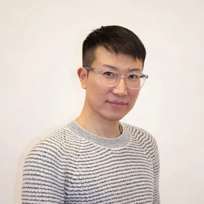 A young man with short dark hair and clear-framed glasses gives a subtle smile while wearing a light-colored crewneck sweater with thin blue horizontal stripes against a solid white background.