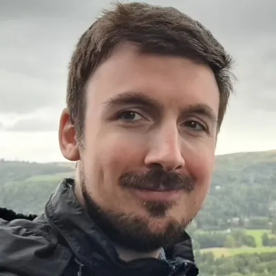 Portrait of a smiling man with dark hair and a beard, wearing a black jacket. He stands outdoors with a scenic, cloudy landscape in the background.