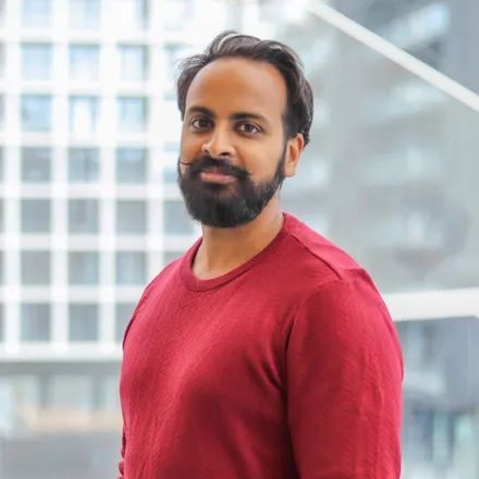 A man with a beard and mustache, wearing a red sweater, stands in front of a modern geometric window.