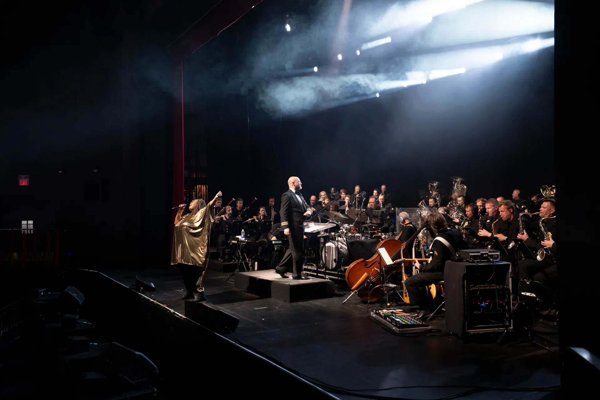 A vocalist in gold and a full orchestra performing under haze and stage beams at Chrysalis