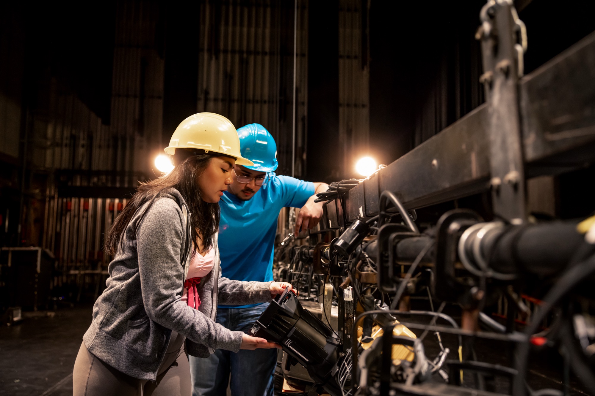 Two students in hard hats hanging a lighting fixture on a stage bar